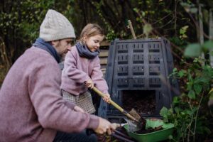 Girl helping father to remove compost from a composter in garden. Concept of composting and